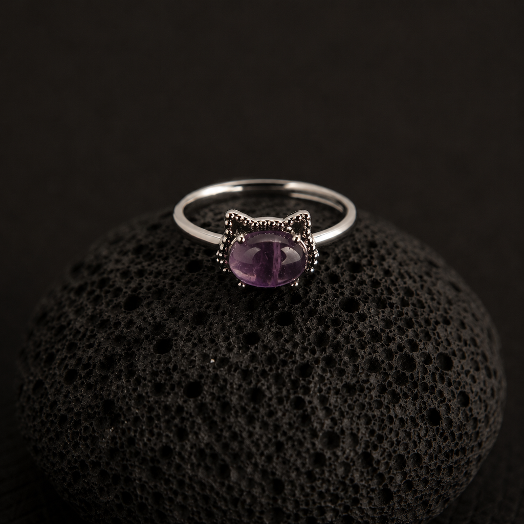 A silver cat-shaped ring with a purple oval gemstone, displayed on a textured black volcanic rock against a dark background.
