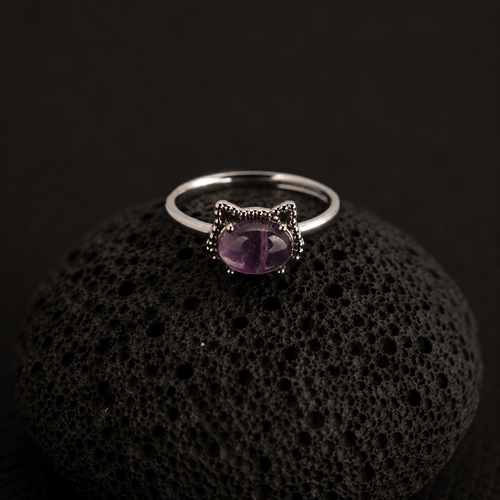 A silver cat-shaped ring with a purple oval gemstone, displayed on a textured black volcanic rock against a dark background.