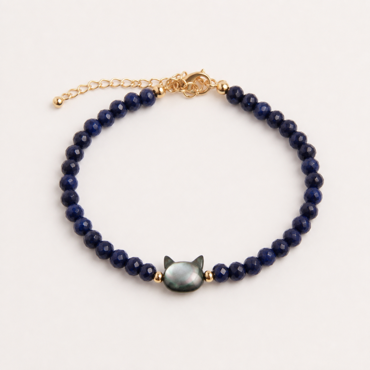 A beaded bracelet featuring dark blue faceted beads, a central iridescent green cat-shaped bead, and gold-toned metal accents including a chain extender and clasp, shown on a light cream background.