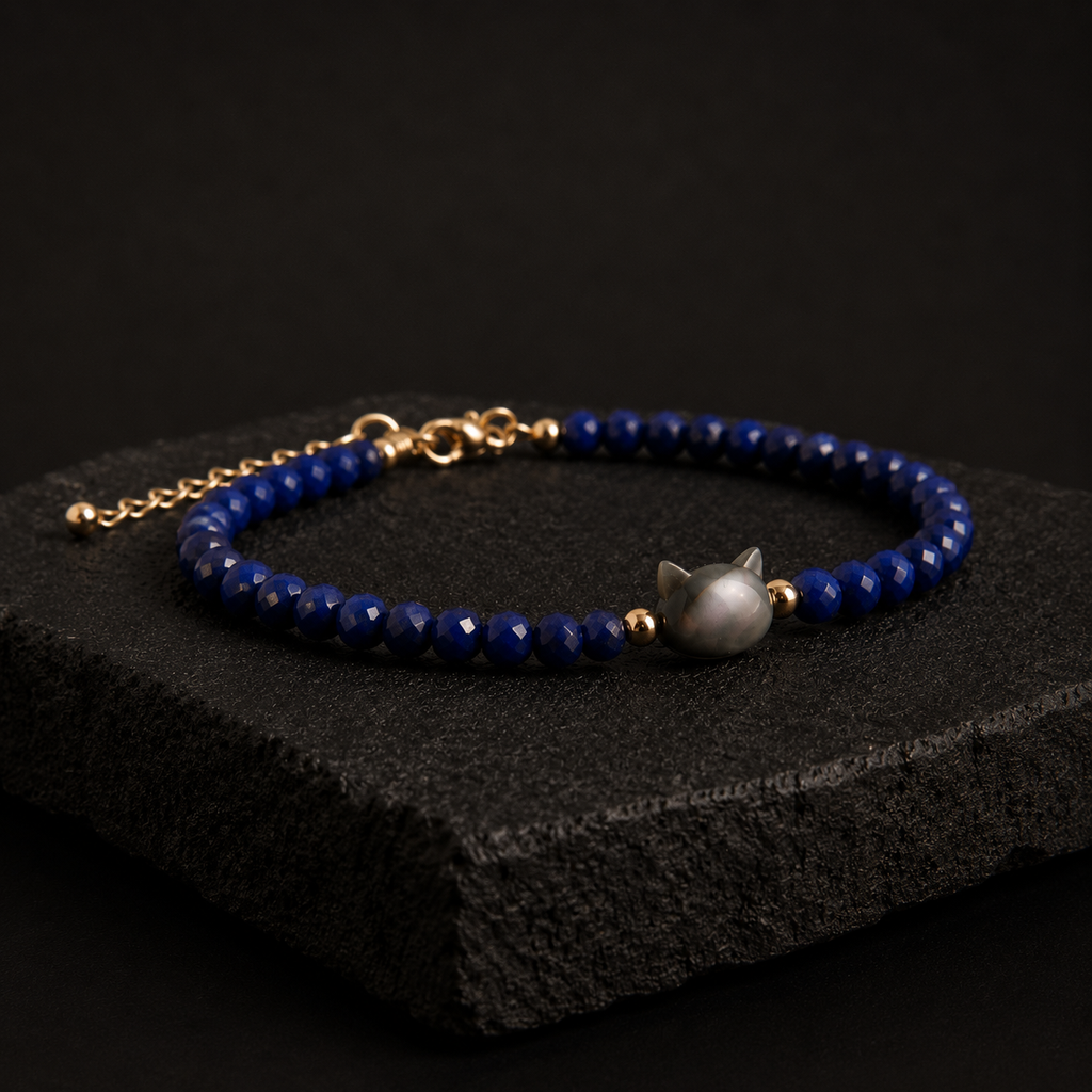 A beaded bracelet with deep blue faceted beads, gold-toned chain and connectors, and a central gray cat-shaped bead, displayed on a dark textured stone block against a black background.