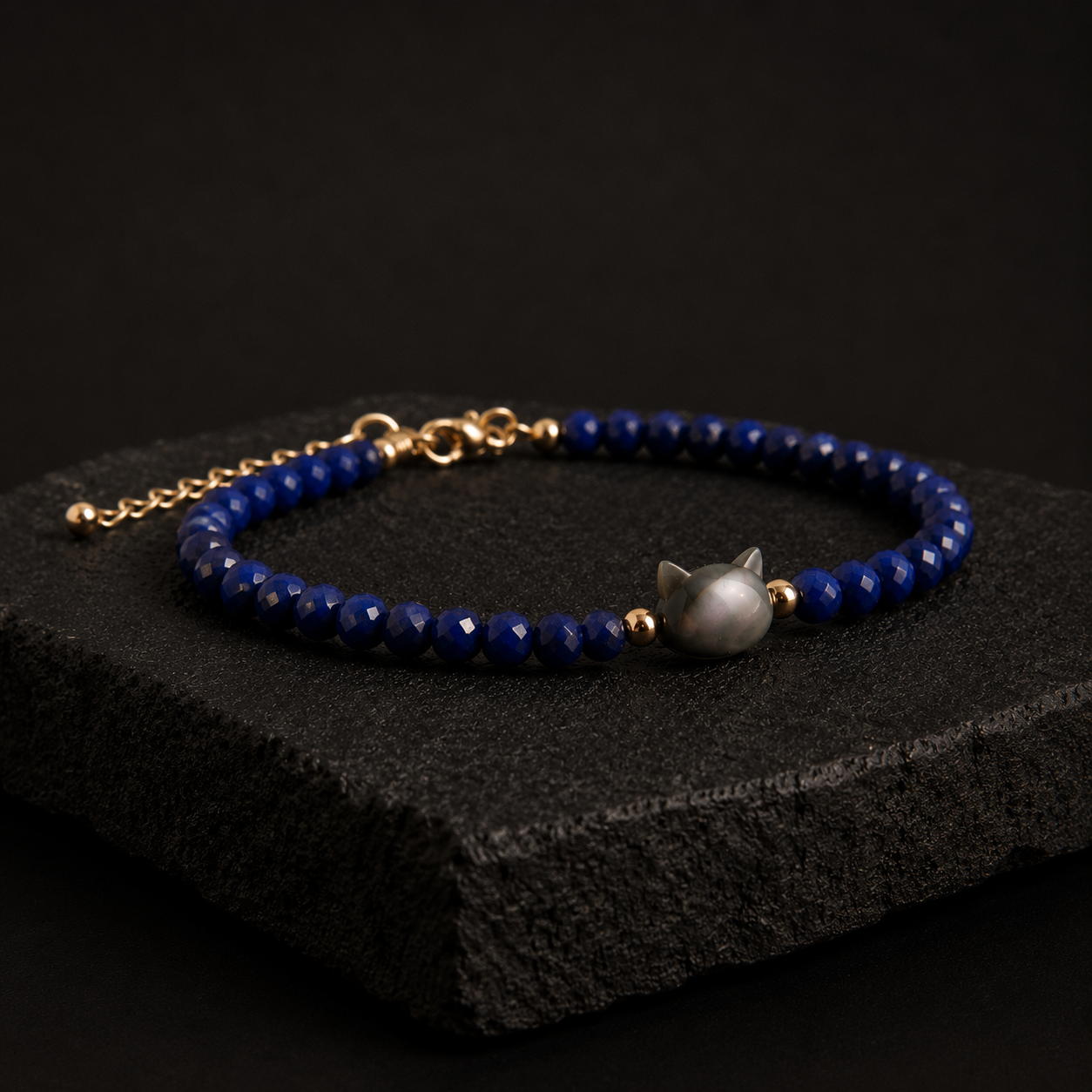 A beaded bracelet with deep blue faceted beads, gold-toned chain and connectors, and a central gray cat-shaped bead, displayed on a dark textured stone block against a black background.