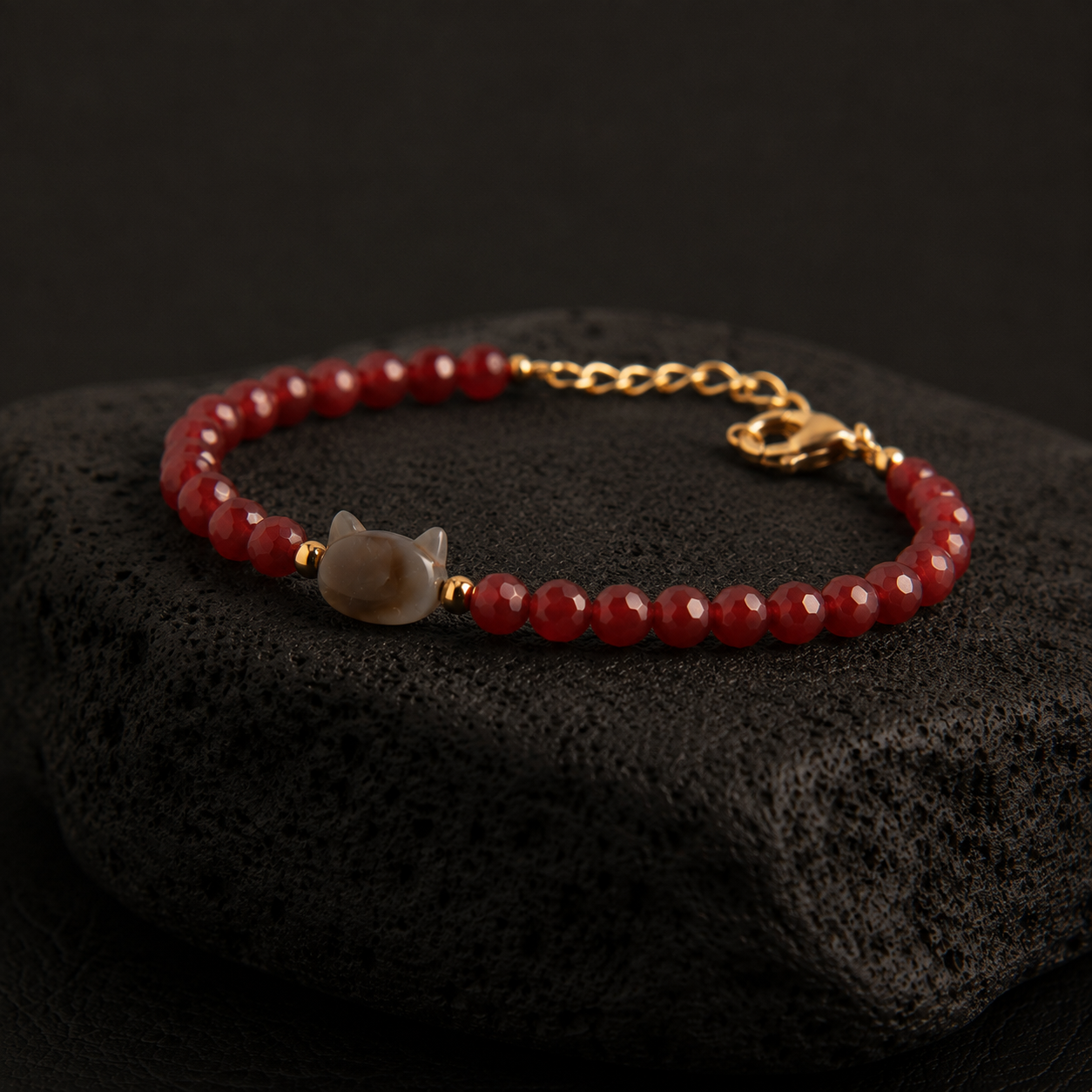 Red faceted bead bracelet with a cat charm and gold accents displayed on a black textured stone.