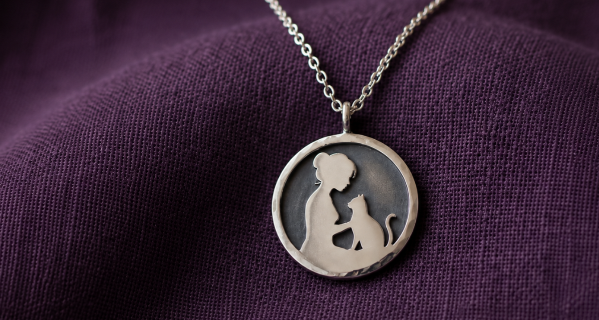 A silver pendant necklace featuring a silhouette of a woman holding a cat, displayed on a textured dark purple fabric background.