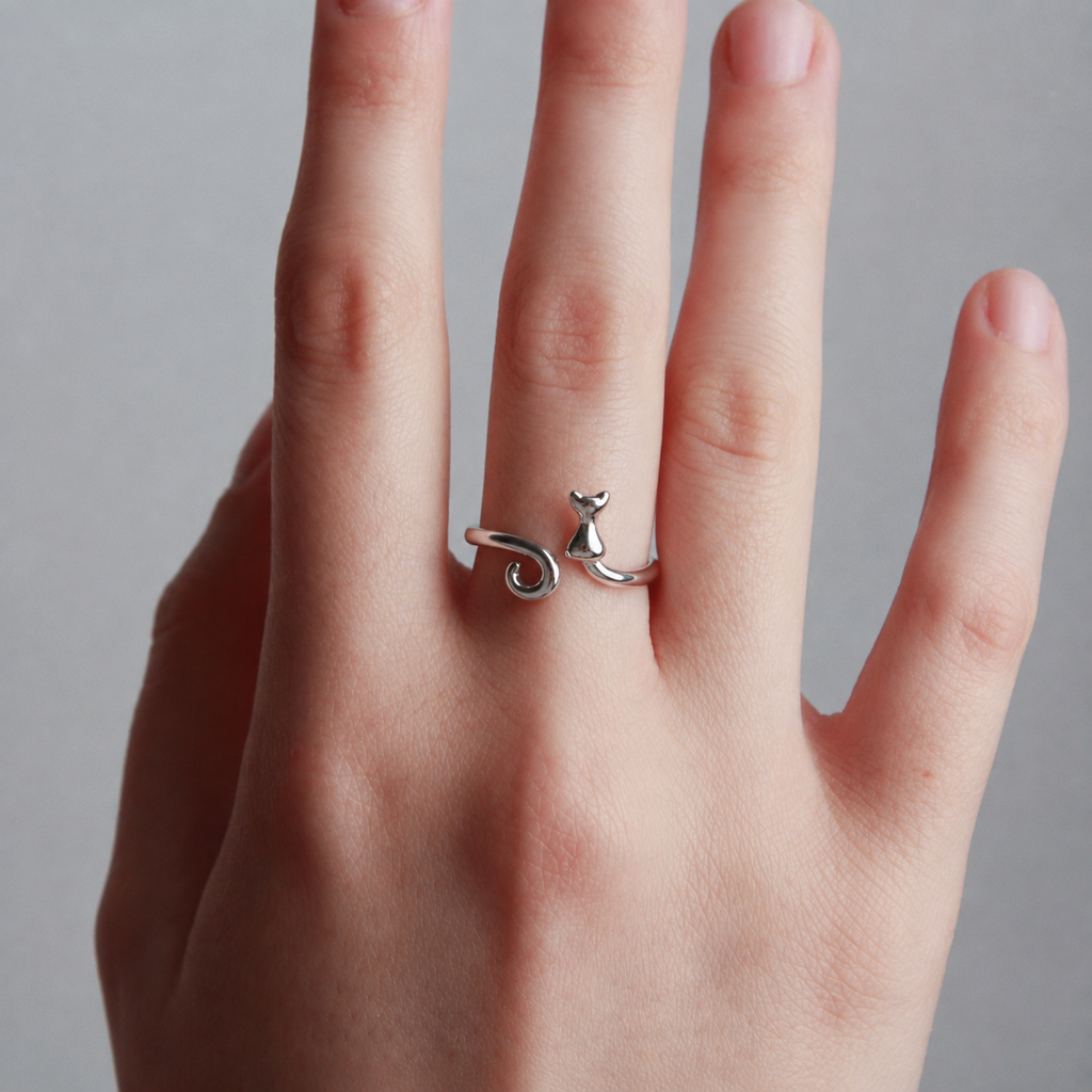 A silver ring shaped like a small cat sitting on a spiral, worn on a person's finger against a neutral gray background.