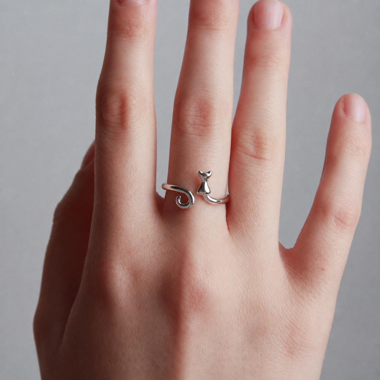 A silver ring shaped like a small cat sitting on a spiral, worn on a person's finger against a neutral gray background.