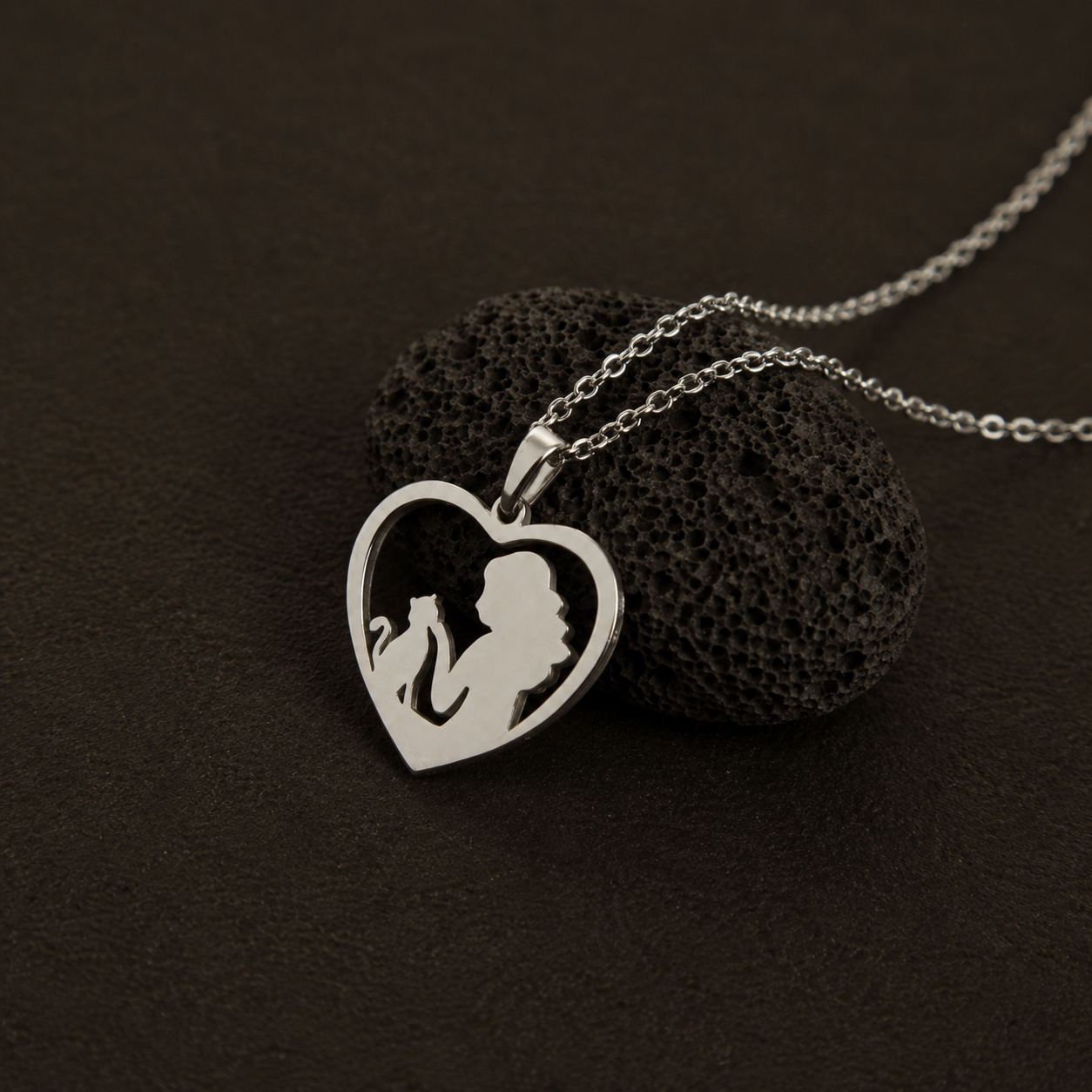 A silver heart-shaped pendant necklace with a silhouette cutout of a woman and cat, resting on a textured black stone against a dark brown background.