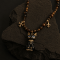 A gothic-style beaded necklace with brown wooden beads, gold-toned accents, and a black cat pendant with blue enamel eyes, displayed on a dark textured rock.