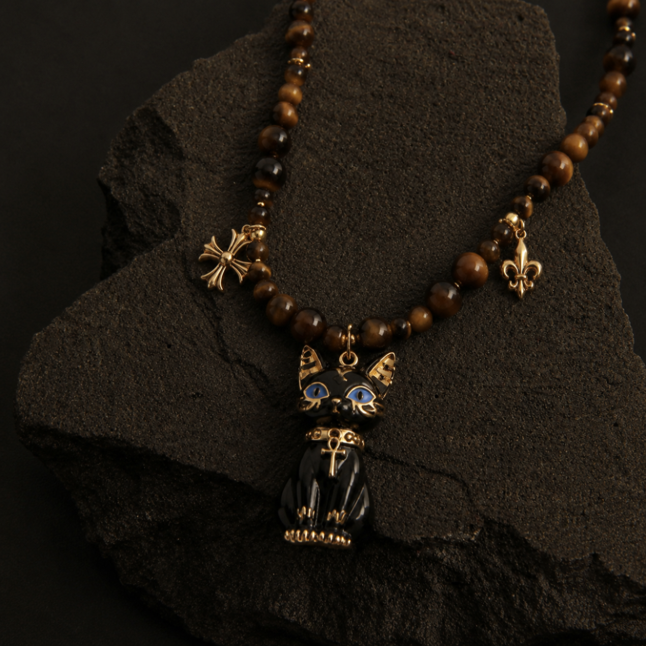A gothic-style beaded necklace with brown wooden beads, gold-toned accents, and a black cat pendant with blue enamel eyes, displayed on a dark textured rock.