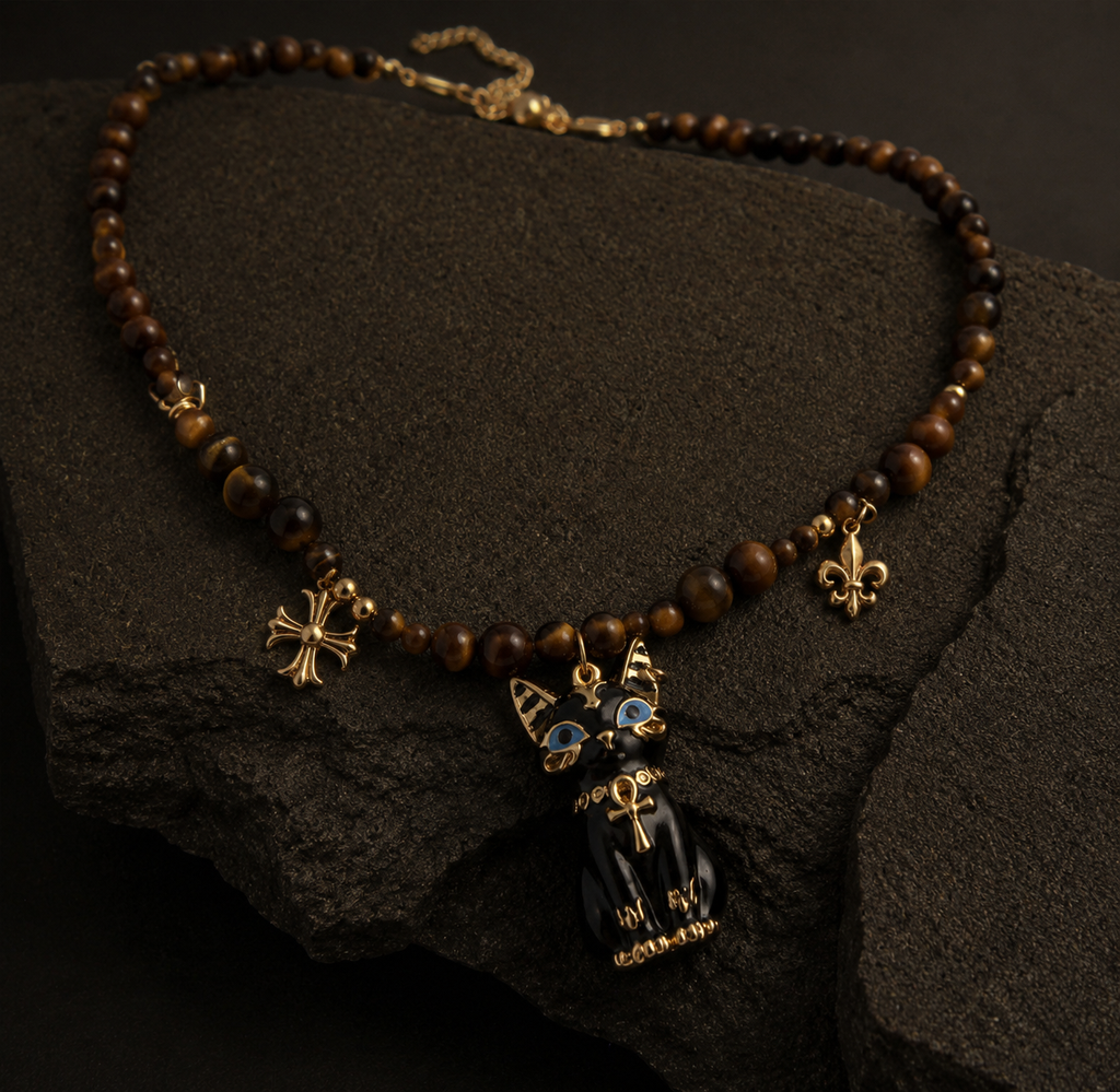 A beaded necklace with brown tiger's eye beads, gold-tone charms including a cross, fleur-de-lis, and a black cat pendant with blue eyes, displayed on a dark textured rock.