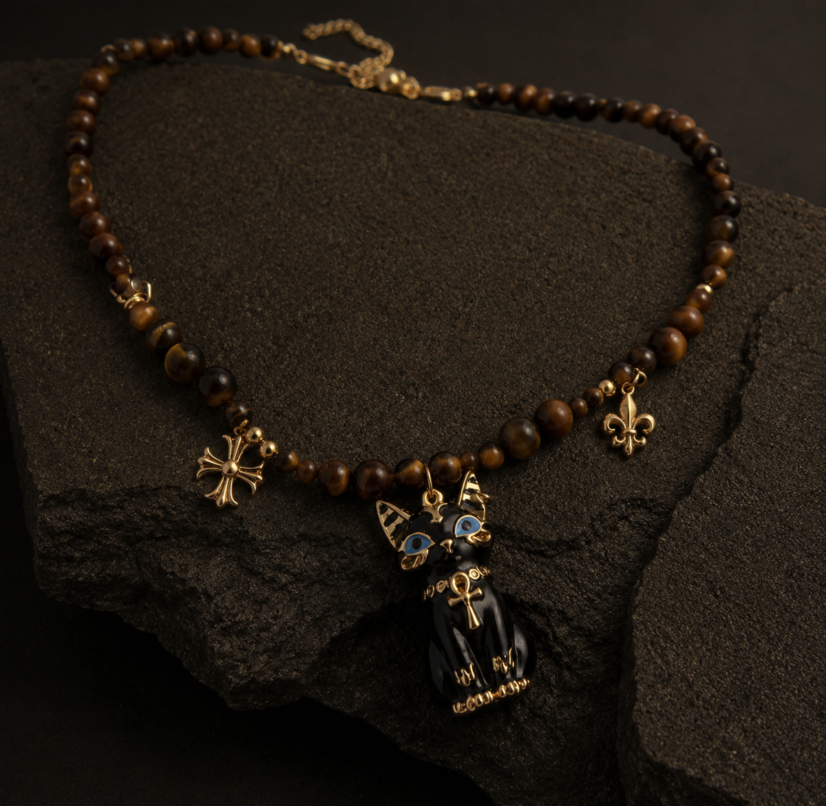 A beaded necklace with brown tiger's eye beads, gold-tone charms including a cross, fleur-de-lis, and a black cat pendant with blue eyes, displayed on a dark textured rock.