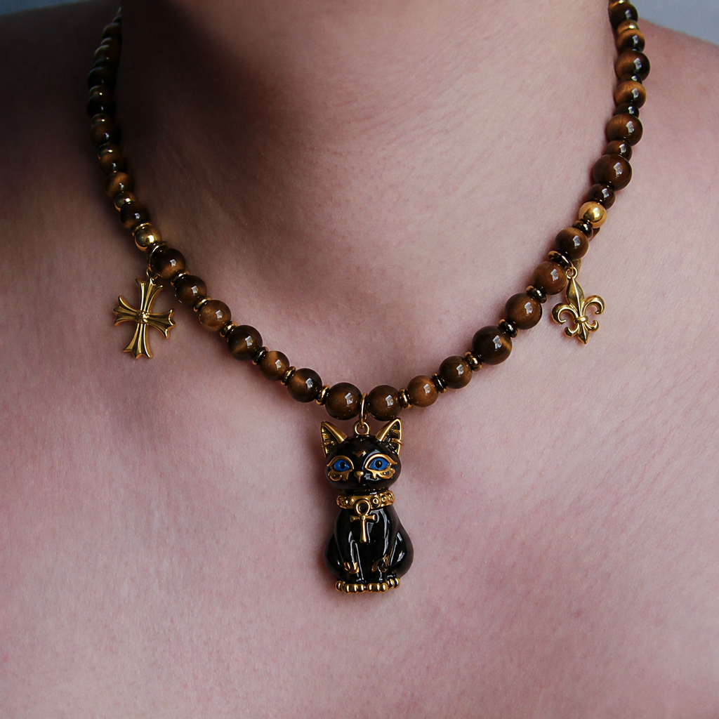A close-up of a person's neck and chest wearing a beaded necklace with a black cat pendant. The necklace features brown and golden-brown beads, a gold cross charm, a gold fleur-de-lis charm, and a central black cat pendant with gold detailing and blue eyes.
