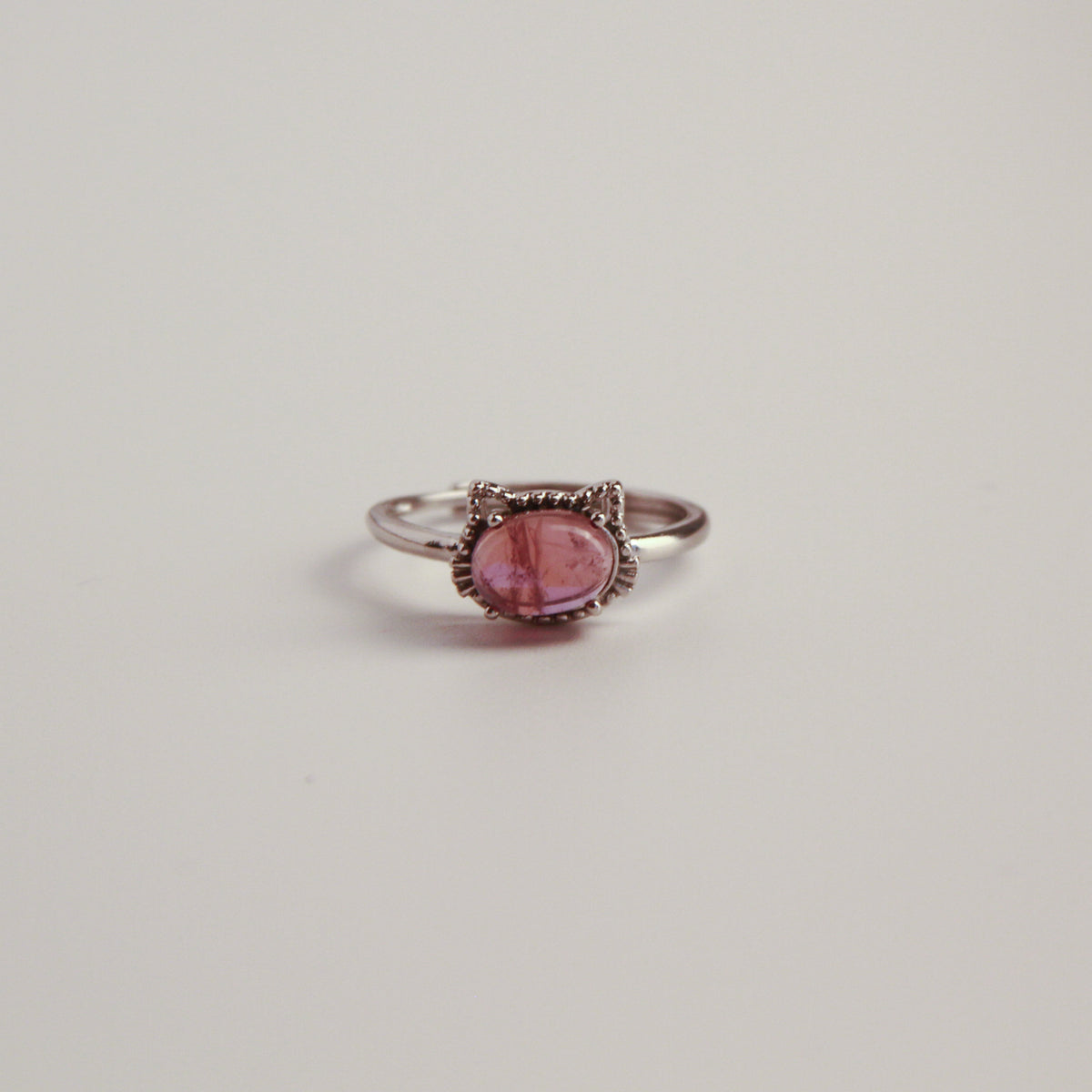 A silver cat ring with an oval pink gemstone in a decorative bezel setting, photographed on a plain light gray background.